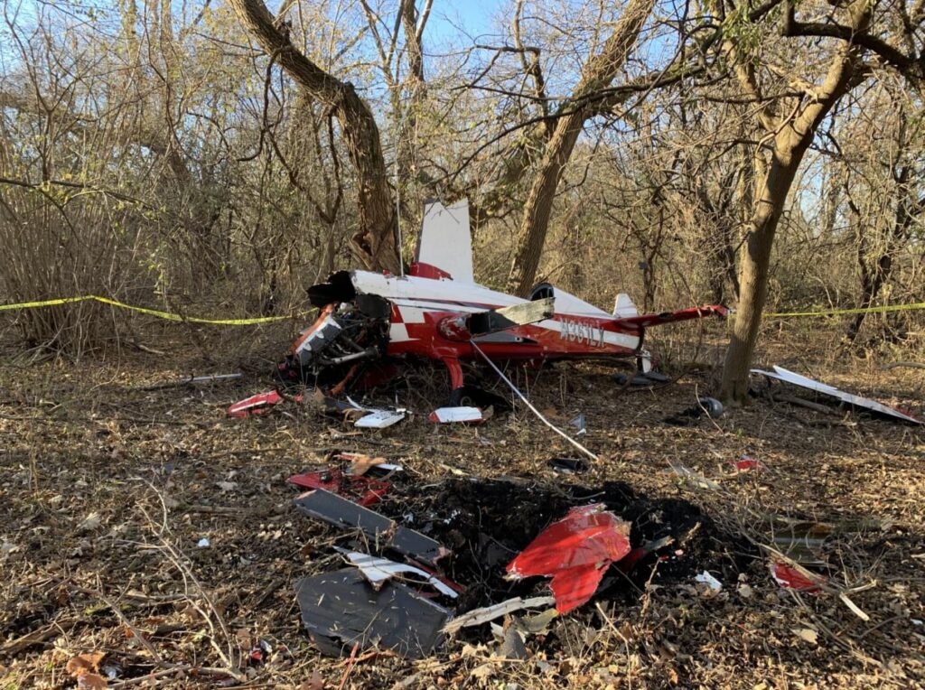 Wreckage of a red and white Extra EA300/LC airplane lying in a wooded area with broken wings and debris scattered across the ground; yellow caution tape is visible in the background.