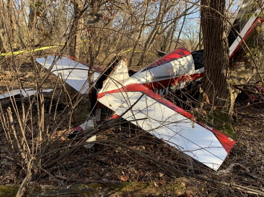 Rear view of the Extra EA300/LC wreckage showing the damaged tail section and horizontal stabilizer wedged against trees in a forested area.