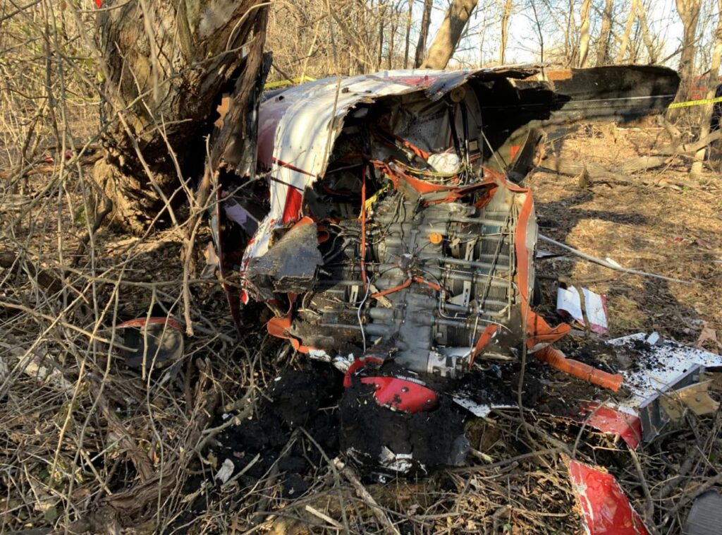 Close-up of the exposed Lycoming AEIO-580 engine from the Extra EA300/LC wreckage, showing significant impact and fire damage with soil and debris surrounding it.