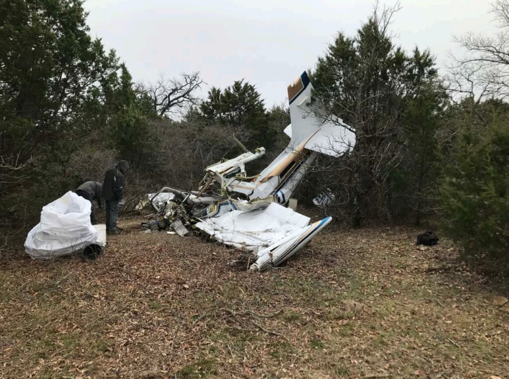Investigators examine the wreckage of a Beechcraft F33A Bonanza in a wooded clearing. The aircraft is nearly vertical against trees with significant nose and fuselage damage, surrounded by debris and bent metal.