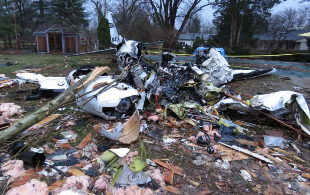 Close-up view of the destroyed fuselage and engine components of the Piper PA-31-350 N400JM resting in a backyard. The wreckage is surrounded by scattered debris, insulation, and tree branches, showing the extensive destruction caused by the crash and post-impact fire.