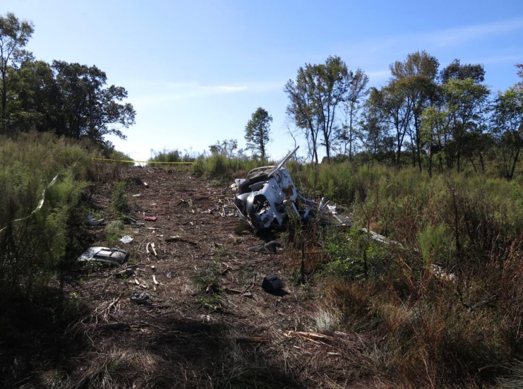 Wreckage trail leading uphill through a cleared, brushy area, with scattered helicopter debris along the ground and the main fuselage lying on its right side near the top of the slope. Trees line both sides under a clear blue sky.