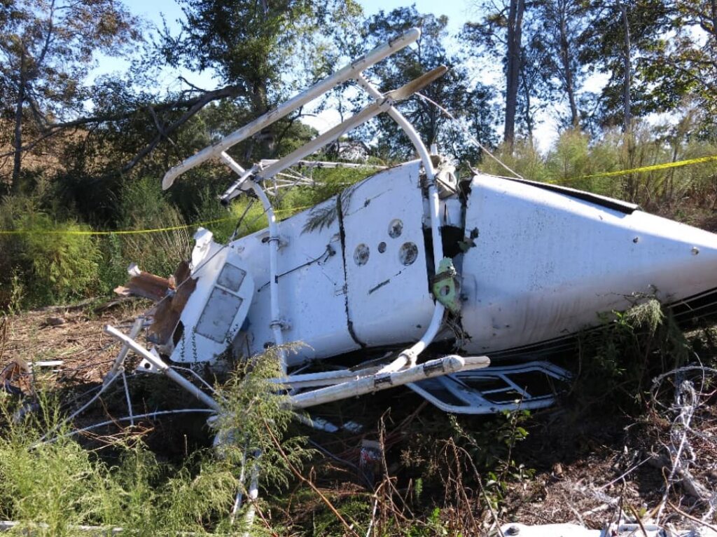 Side view of the overturned helicopter fuselage resting in vegetation, with significant impact damage, broken framing, and detached components visible around the wreckage area.