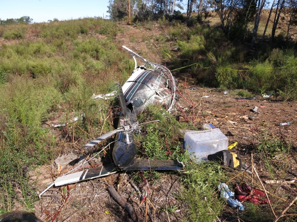 Tail boom and tail rotor assembly lying separated from the main fuselage in a brush-covered clearing, with debris and broken components scattered around the impact site.