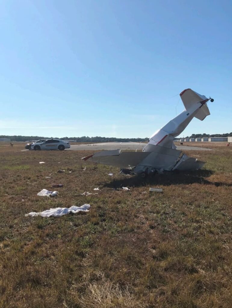The same crashed Bushby Mustang II aircraft is shown from a rear angle, still resting nose-down in the grass near a runway. Debris is scattered across the field, and a sheriff’s vehicle is parked on the pavement in the background under a clear sky.