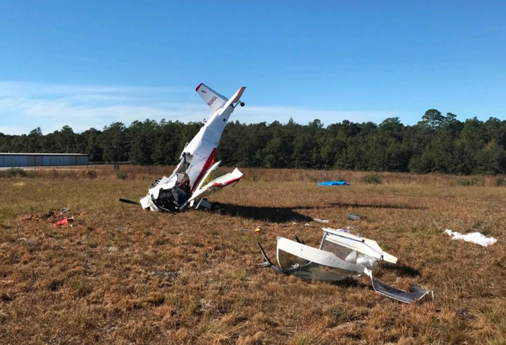 A wide view of the accident site shows the damaged Bushby Mustang II in the center with its tail pointed upward. Additional debris, including a separated canopy piece, is scattered across the grass. Trees line the background beneath a clear blue sky.