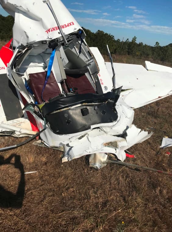 A close-up view of the crushed cockpit area of the Bushby Mustang II shows the severely deformed nose section, exposed seat cushions, and partially detached components. The surrounding grass is flattened, and a shadow of the photographer is visible in the foreground.