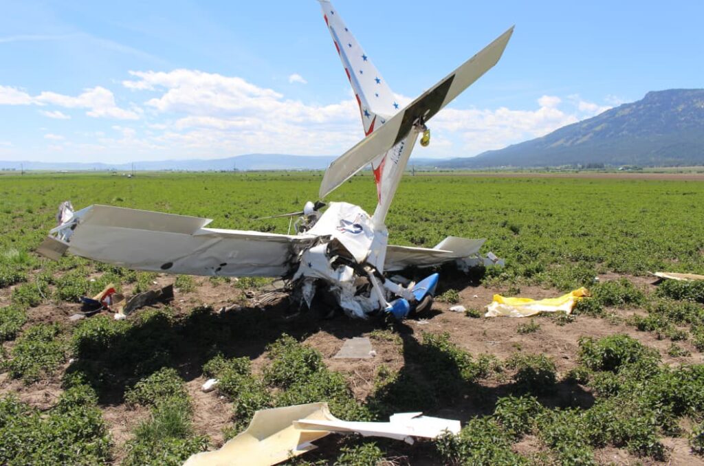 Rear view of the crashed Titan II aircraft in a flat farm field, with the tail standing nearly vertical and the fuselage heavily compacted at the point of impact. Both wings are crumpled, and debris is scattered around the site. Rolling hills and distant mountains appear under bright daylight.