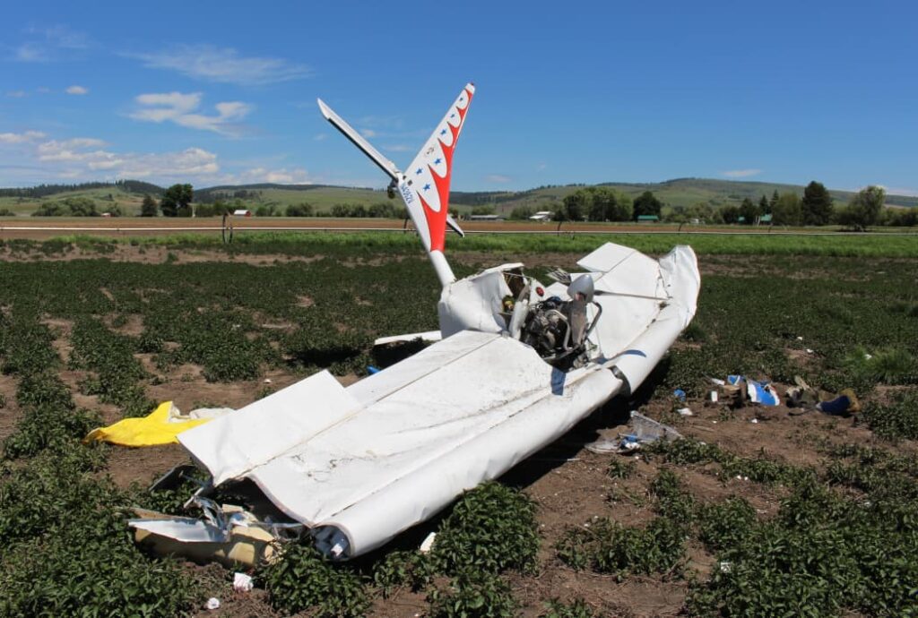 Side view of the Titan II aircraft wreckage resting in a field, showing a crushed fuselage, twisted wings, and the partially detached engine mounted forward. The tail section marked N4362V remains upright. The surrounding farmland and distant hills are visible beneath a sunny, blue sky.