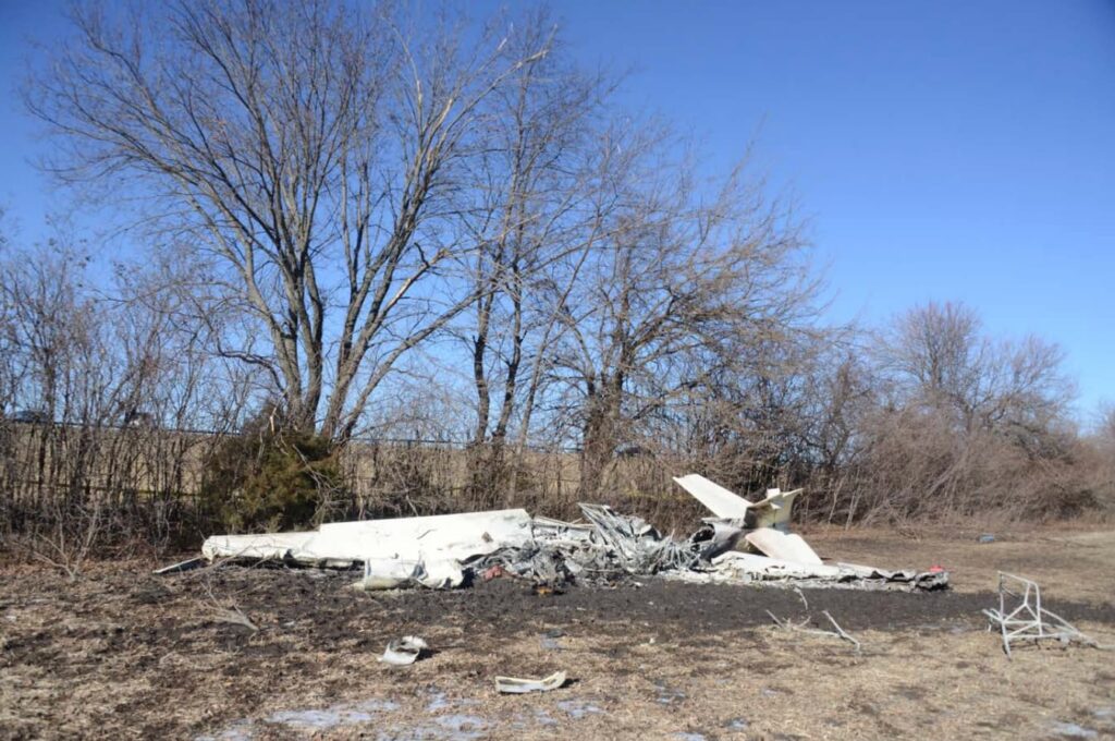 Wide view of the crash site showing the Mooney M20S wreckage resting in a clearing beside leafless trees. The aircraft is severely burned, with the fuselage collapsed and reduced to metal debris. The tail assembly remains recognizable but charred. Surrounding ground is blackened from fire.