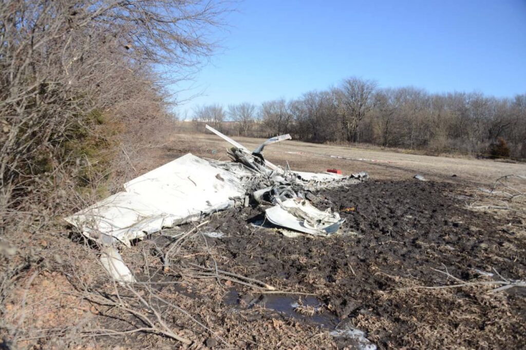 Closer angled view of the destroyed Mooney M20S wreckage lying on burned, muddy ground. The fuselage and wings are crumpled and heavily fire-damaged, with twisted airframe components scattered around. Trees and open terrain are visible in the background.