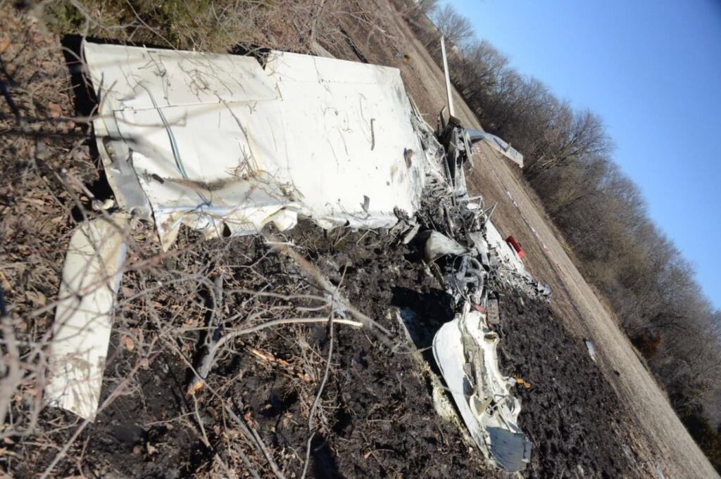 Side view of the Mooney M20S crash site showing a long trail of wreckage. Wings and fuselage sections are flattened and melted, with debris extending toward an open field. The ground is churned and darkened from impact and fire.