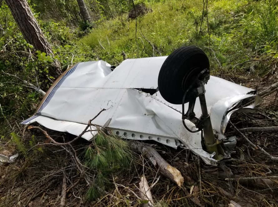 A separated section of an aircraft wing lies in a forested area. The wing is crumpled and bent, with the landing gear still attached and pointing upward. Broken tree branches and scattered debris are visible among the underbrush.