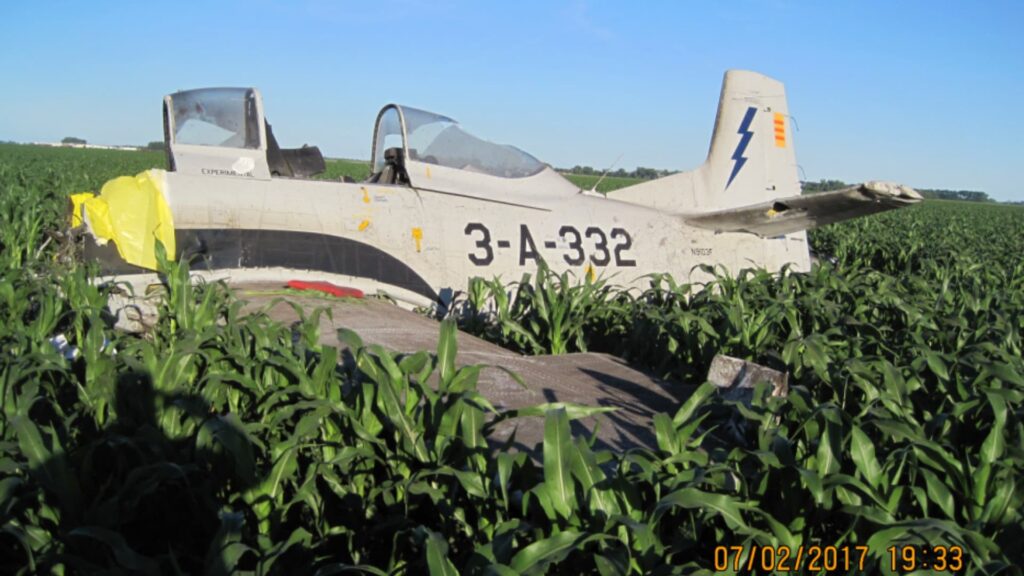 Wreckage of a North American T-28 aircraft resting in a cornfield, viewed from the left side, with the fuselage largely intact, canopy sections visible, and yellow material covering damage near the nose under clear blue skies.