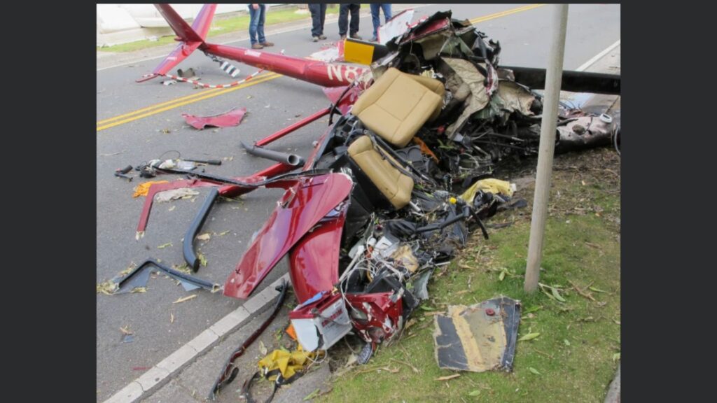 A heavily damaged red Robinson R44 helicopter lies in pieces on a residential street. The fuselage is crushed and torn open, exposing wiring, mechanical components, and the collapsed beige cabin seats. Sections of rotor blades and airframe debris are scattered across the roadway and onto the grass. A portion of the tail boom, still intact, extends behind the wreckage. Investigators or first responders stand in the background on the street.