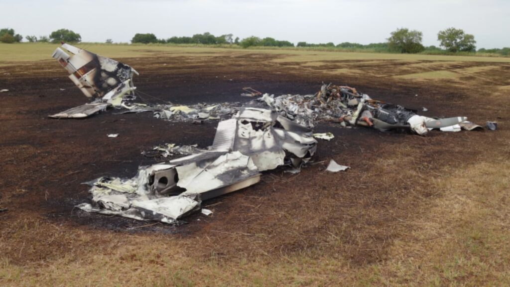 A burned and fragmented Piper PA-34 wreckage lies scattered across a scorched patch of grass. The tail section remains partially recognizable on the left side of the scene, but the fuselage and wings are almost entirely consumed by fire damage, leaving twisted metal and ash. The surrounding field shows a large burned area where the post-impact fire spread.