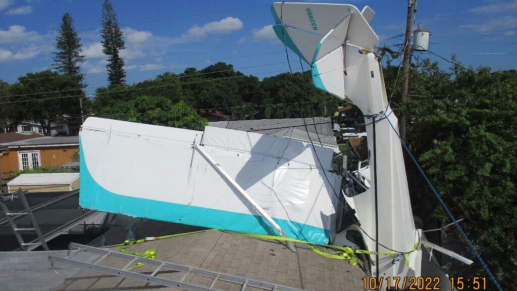 Vertical stabilizer and fuselage section of an experimental airplane resting on a residential rooftop, secured with straps during recovery operations.