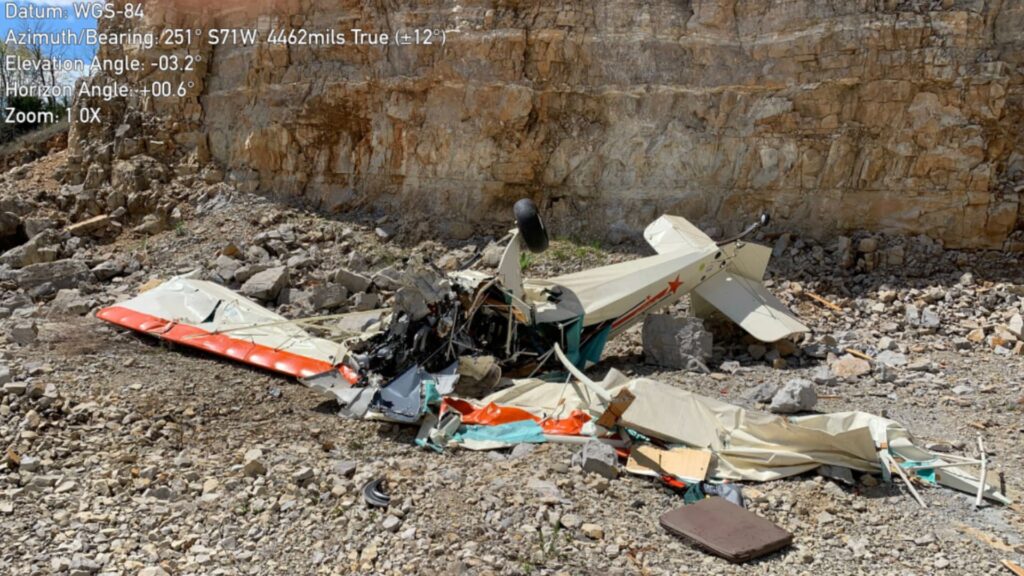 Wreckage of a single-engine light airplane lying on rocky ground at the base of a vertical quarry wall. The aircraft is heavily damaged with the fuselage crushed, wings broken and folded, and debris scattered across loose rock. The tail section remains partially intact, while the engine and cockpit area are destroyed. A sheer rock face rises directly behind the wreckage.
