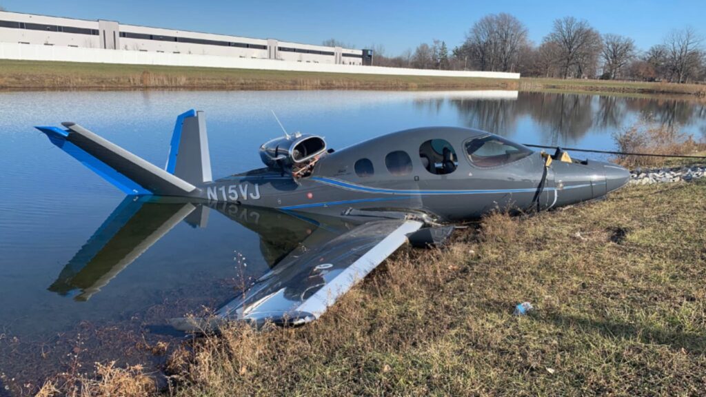 Side view of a Cirrus SF50 Vision Jet, registration N15VJ, stopped at the edge of a retention pond. The left wing and tail are partially submerged, while the fuselage remains intact. The CAPS system has deployed, and the aircraft appears largely undamaged aside from landing impact.
