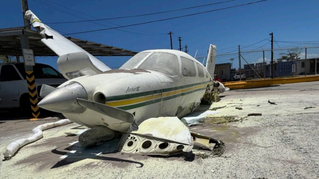 Damaged Piper PA-28 Cherokee resting on a concrete parking area after a crash, with the nose low to the ground, a bent propeller, collapsed landing gear, and visible structural damage to the fuselage and wing under clear daylight conditions.