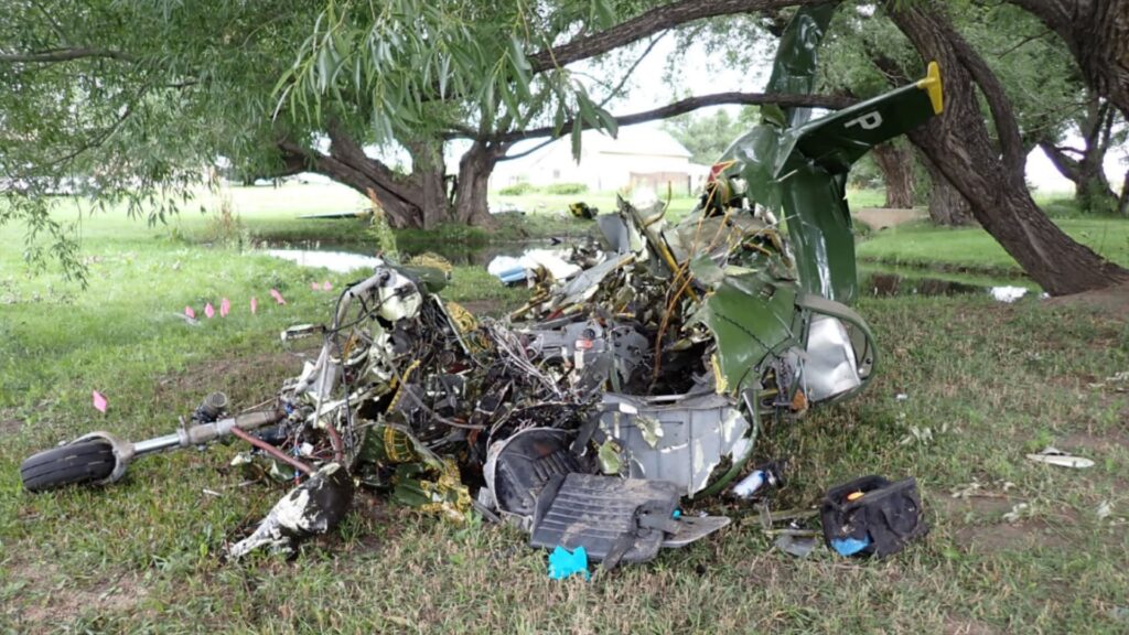 Severely damaged aircraft wreckage resting on grass near trees, with the fuselage crushed and fragmented, debris scattered around the impact site, and a shallow water feature visible in the background.