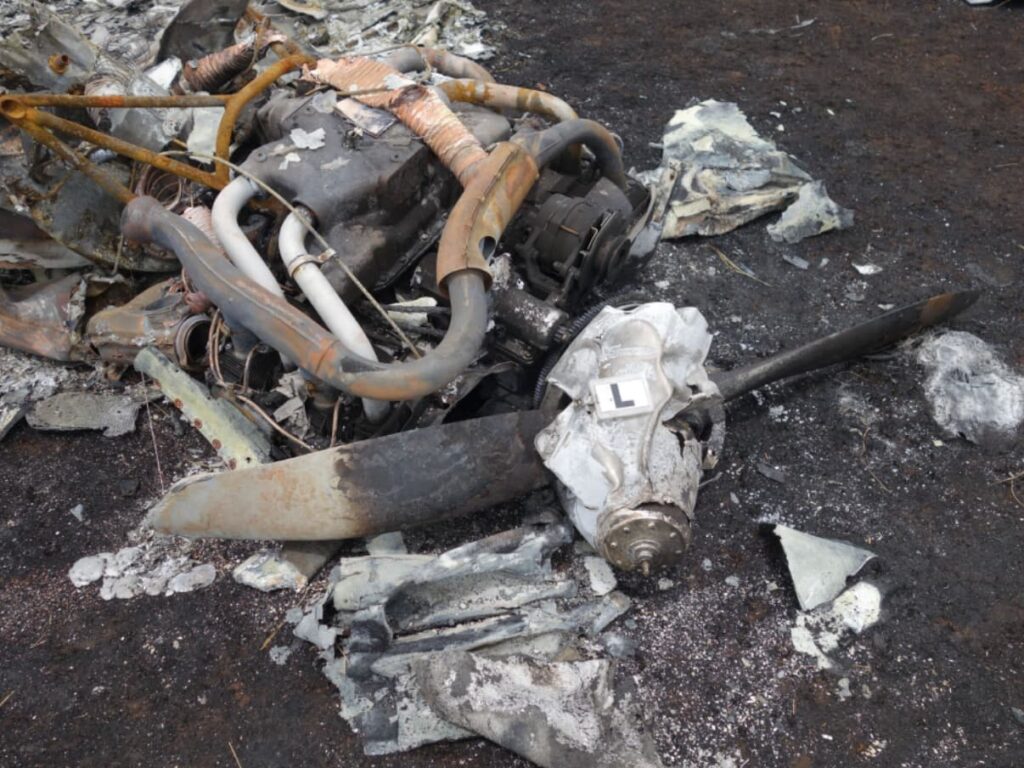 A heavily fire-damaged engine from the left side of the Piper PA-34 rests among charred debris. The propeller hub is attached but the blades show little rotational scoring, consistent with low or no power at impact. Burned and melted components surround the engine, and a label marked “L” is placed atop the propeller hub for identification.