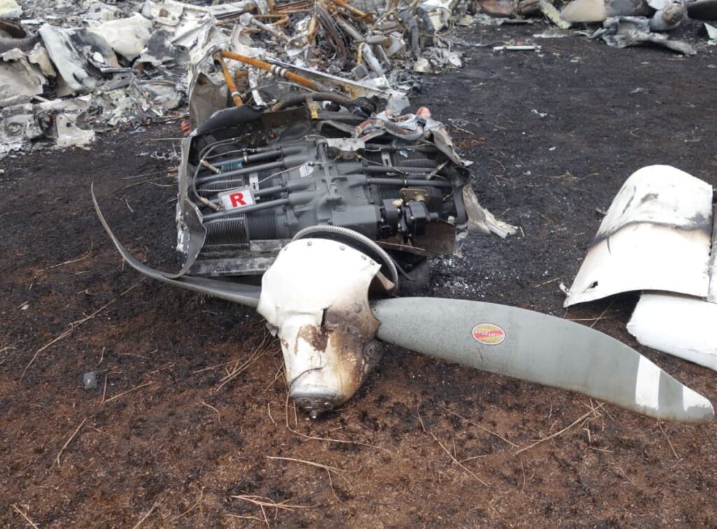 The right engine from the Piper PA-34 lies separated from the fuselage, showing significant fire damage but intact cylinders and structure. The propeller remains attached and is bent and scarred, with clear indications of rotation at impact. A tag marked “R” identifies the engine. Burned ground and additional wreckage pieces are visible around it.
