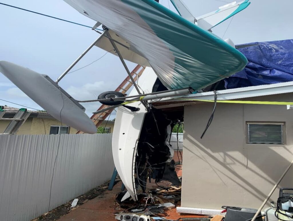 Wreckage of an experimental airplane embedded in a residential structure, with the fuselage nose-down against a wall and the wing resting on the roof.