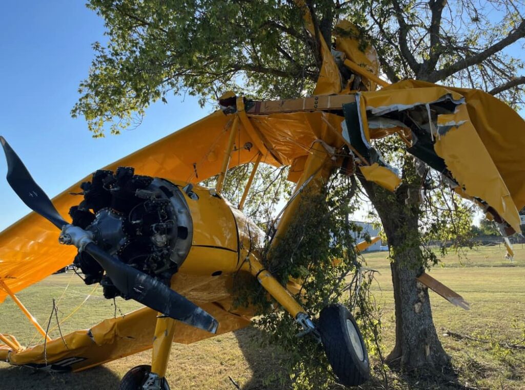 Close-up view of the damaged yellow aircraft showing the exposed radial engine and bent propeller, with the wing structure shredded and lodged in a tree following a crash on a grassy area.
