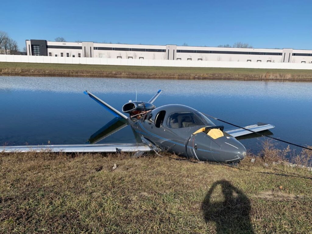 Cirrus SF50 Vision Jet resting partially in a retention pond after CAPS deployment. The airplane is upright with the nose on the grassy bank and the wings extending into the water. The parachute has been cut away, and the engine nacelle on top of the fuselage is exposed. Industrial buildings and a clear blue sky are visible in the background.