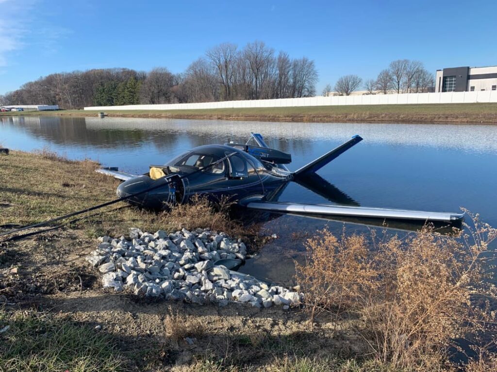 Front-left angle view of a Cirrus SF50 Vision Jet following a parachute landing into a retention pond. The nose rests on the shoreline rocks and grass, with both wings extending into the water. Recovery lines are attached to the fuselage, and the aircraft remains upright under clear daylight conditions.