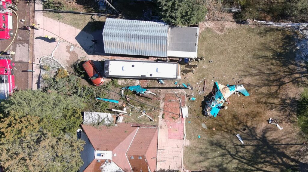 Overhead aerial view of an airplane crash site in a residential area, showing scattered turquoise aircraft debris across a yard, a damaged structure, emergency vehicles on a nearby street, responders on scene, and the main wreckage separated from the wings.
