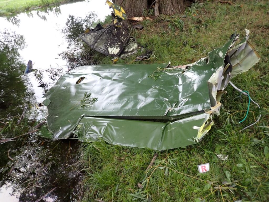 Crumpled green aircraft wing section lying partially beside a small pond, showing extensive deformation, torn metal edges, and impact damage.