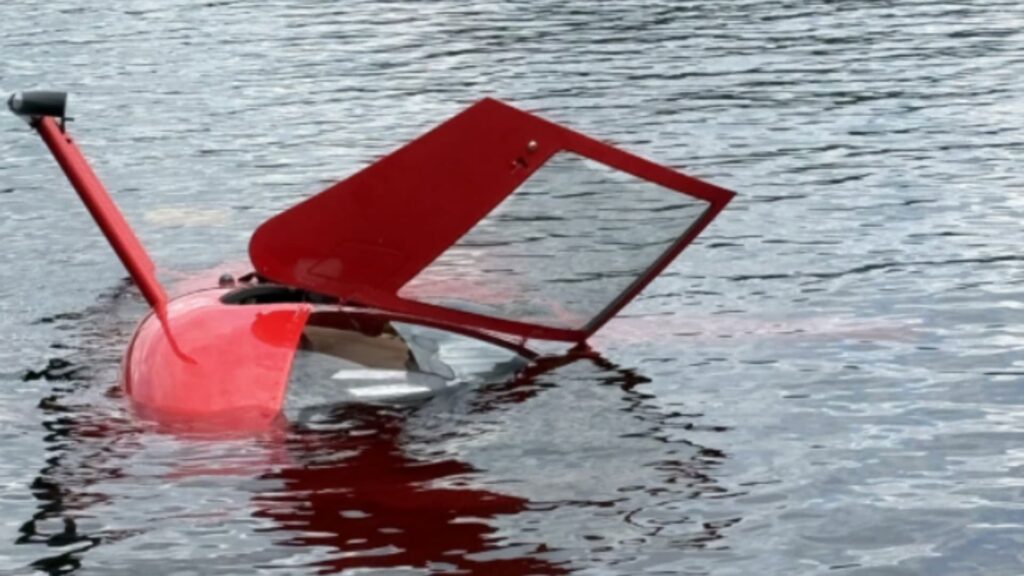 Partially submerged red helicopter resting in calm water, with the cabin and open door visible above the surface after a water impact.