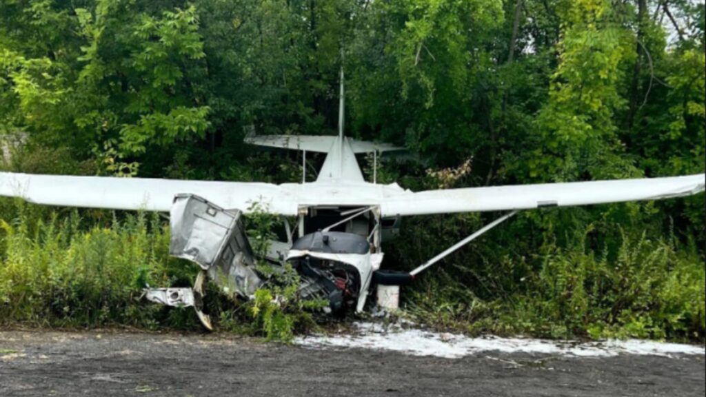 Front view of a damaged Cessna 172 stopped at the edge of a wooded area, showing a crushed nose section and bent wings after a forced landing.