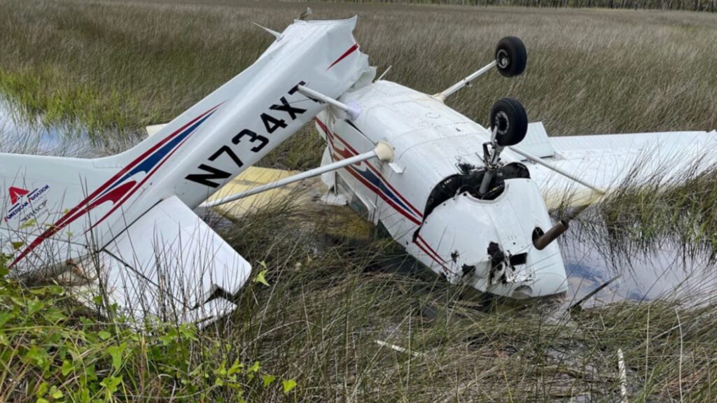 Cessna 172 N734XT resting upside down in a grassy marsh area after a forced landing, with landing gear in the air and visible damage to the fuselage and wings.