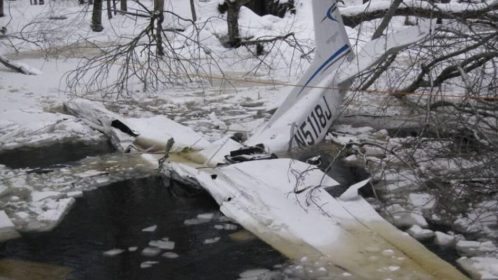 Wreckage of a Cessna 172S resting partially submerged in an ice-covered river, with the tail section upright and wings broken and surrounded by floating ice.