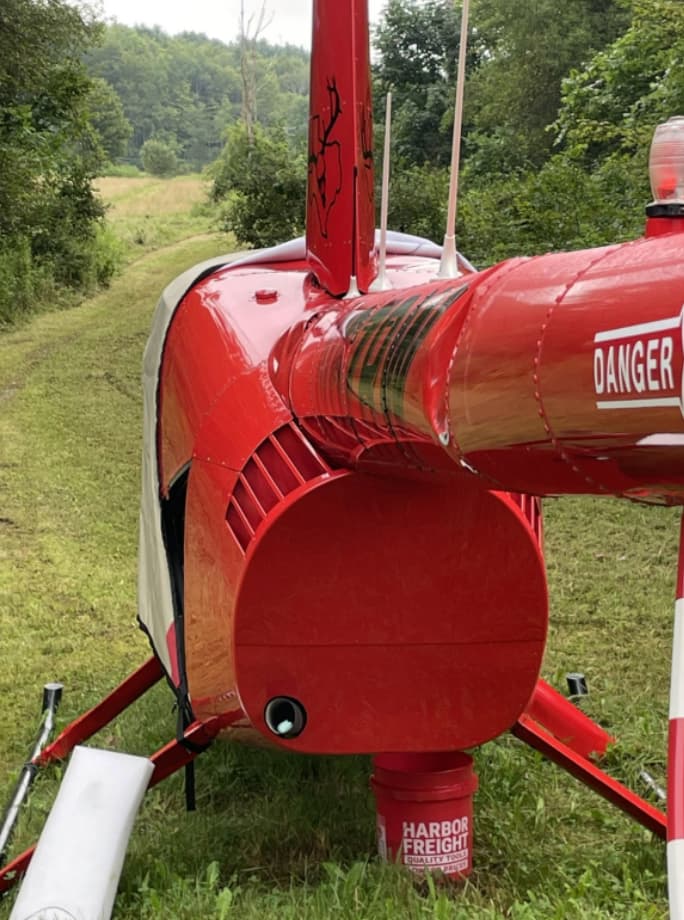 Rear view of a red helicopter parked on grass, showing the tail boom and tail rotor assembly with visible structural damage near the tail section.