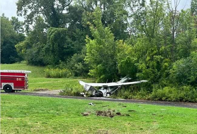 Cessna 172 resting off a paved path after an emergency landing, with visible ground scars in the grass leading to the aircraft and a fire truck parked nearby.