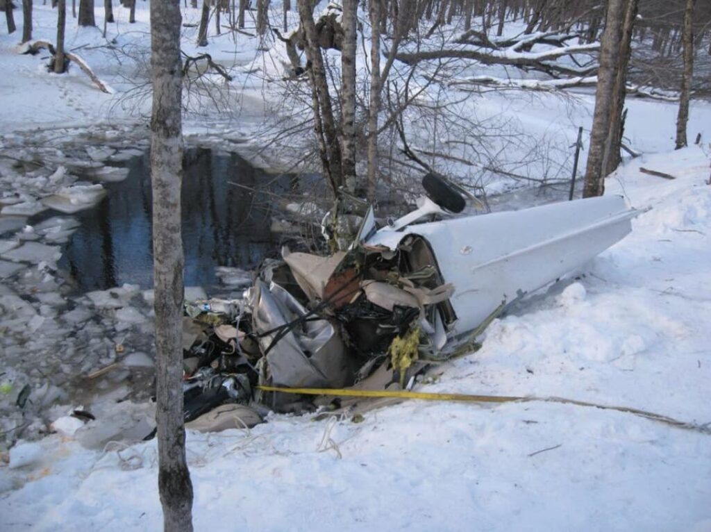 Crushed fuselage of a Cessna 172S lying on a snowy riverbank beside an ice-filled stream, with trees surrounding the accident site.