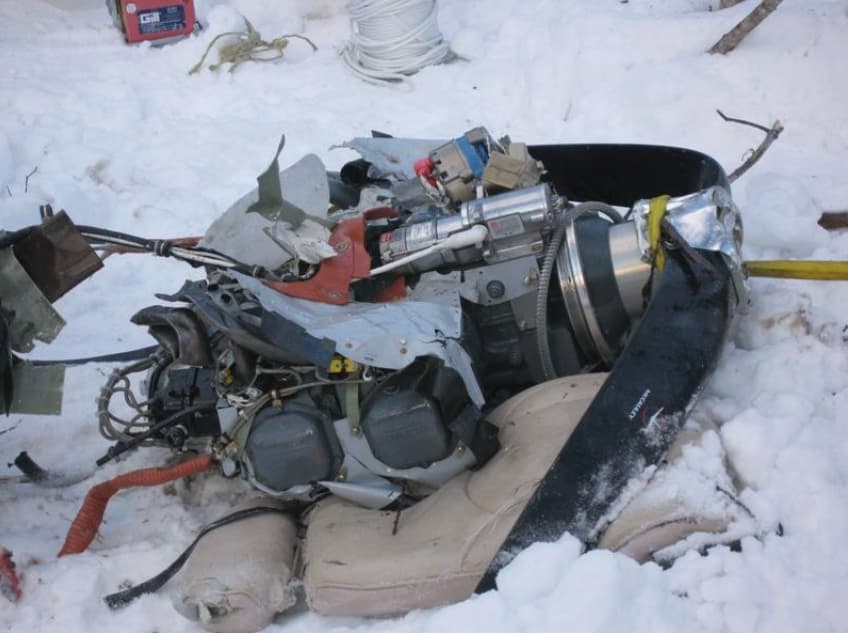 Aircraft engine and bent propeller separated from the airframe, resting on snow after impact, showing extensive impact damage.