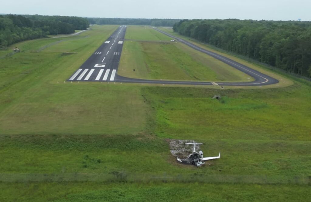 Aerial view of a 5,000-foot asphalt runway surrounded by grass and trees, with a crashed HondaJet resting beyond the runway end in a grassy embankment.