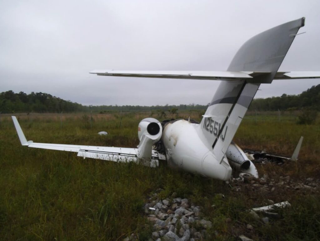 Rear-left view of a damaged HondaJet (N255HJ) resting in tall grass and rocks beyond the runway, showing fire damage to the fuselage and right engine area.