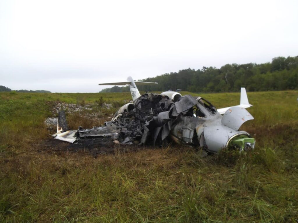 Side view of the heavily burned HondaJet fuselage in a grassy area, with the cockpit and center section destroyed by fire and debris scattered nearby.
