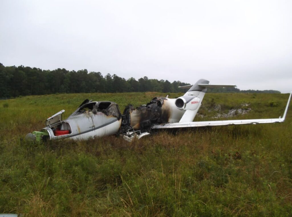 Opposite side view of the wrecked HondaJet showing severe fire damage to the cabin and center fuselage, the tail and wings partially intact, resting in tall grass beyond the runway.