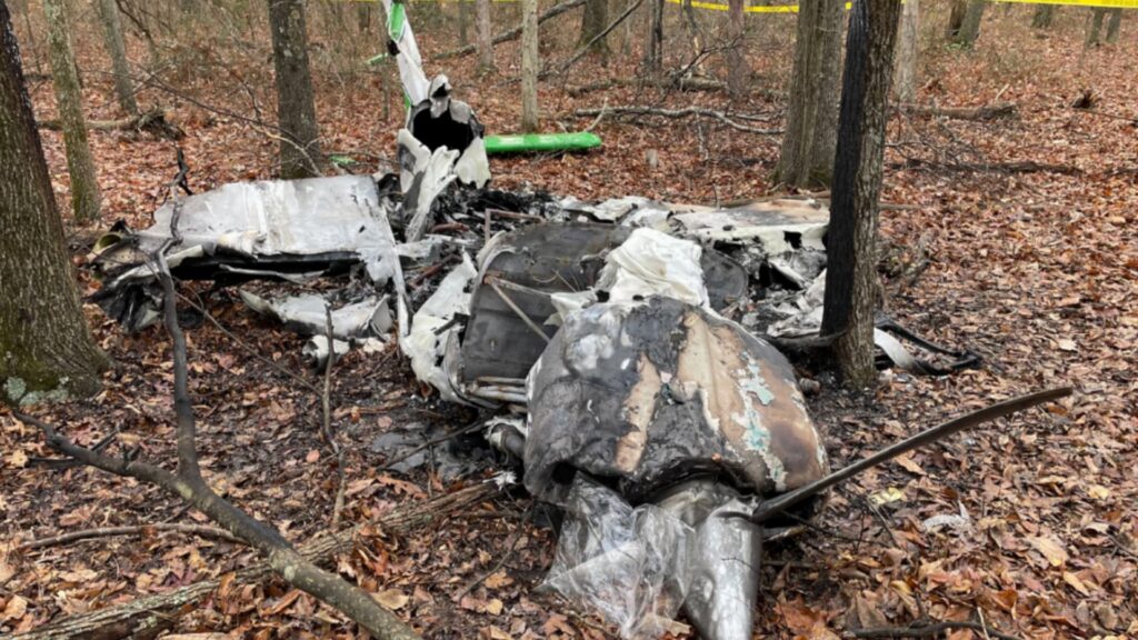 Wreckage of Beech C23 aircraft scattered in a wooded area, showing burned and fragmented fuselage and engine components among trees with fallen leaves and caution tape marking the crash site.