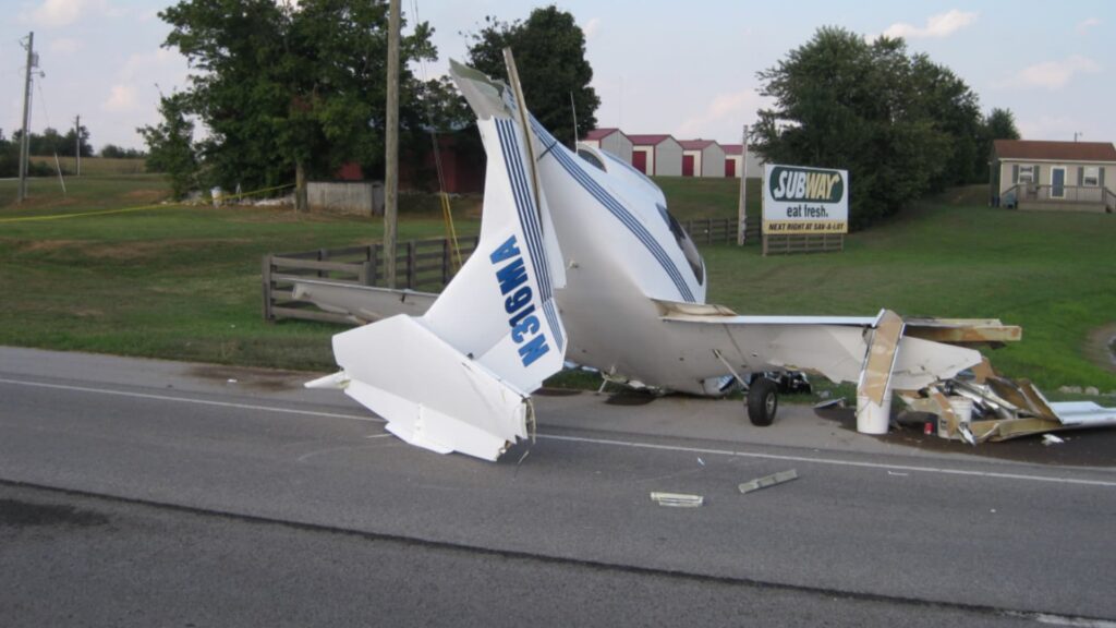 A heavily damaged white Diamond DA40 aircraft rests nose-down along the edge of a paved road. The tail section is tilted sharply upward, and the registration number “N316MA” is visible on the vertical stabilizer. The wings are broken and partially detached. Debris is scattered on the roadway and nearby grass. In the background are utility poles, trees, a small building, and a roadside Subway restaurant sign.