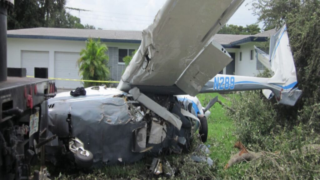 Wreckage of Cessna T337G (N289) after a crash in a residential yard, showing severe nose and fuselage damage, bent wing, and debris scattered near a house and tow truck.