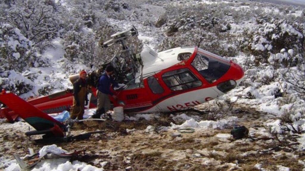 A red and white Eurocopter AS350 B3 helicopter rests on its right side in snowy, brush-covered terrain. The main rotor assembly is damaged and exposed, and the fuselage is tilted downhill on uneven ground. Two individuals wearing winter clothing stand beside the cockpit area. Snow covers the surrounding desert vegetation and hills in the background.