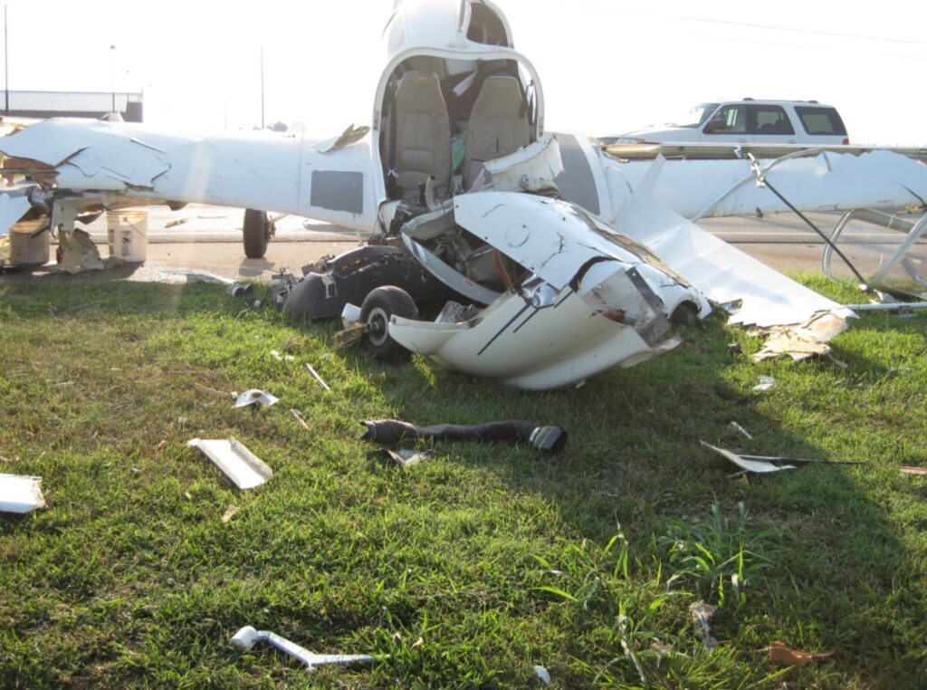 Front view of the crashed Diamond DA40 showing severe structural damage to the nose and cockpit area. The engine cowling is crushed and torn open, with internal components exposed. The cabin door is open, revealing the two front seats inside. Pieces of the aircraft, including sections of the wing and fuselage, are scattered across the grass near a paved surface with a parked SUV in the background.